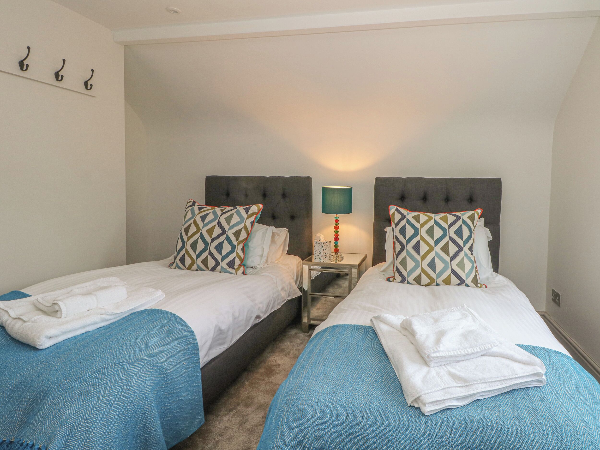 A hotel room with two beds, each with a blue herringbone bedspread, white linens, and patterned pillows. There is a nightstand with a lamp between the beds.