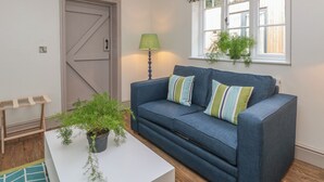 A living room with a blue sofa, a white coffee table, a green plant, and a patterned window valance. - Lakeside Cottage (Macclesfield)