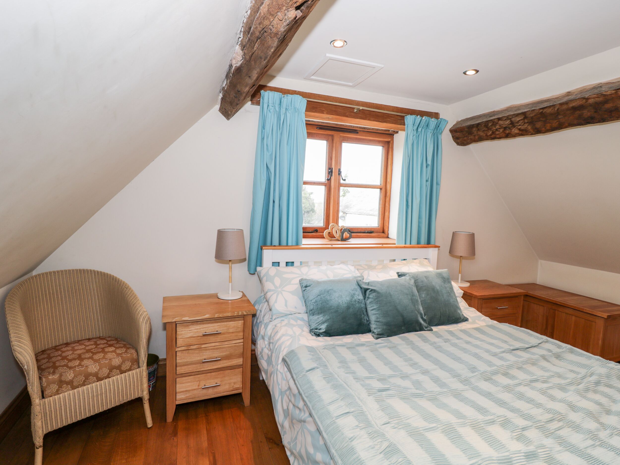 Une chambre avec un plafond en pente, un lit avec une literie à motifs, une chaise en osier, une table de chevet avec des lampes et une fenêtre avec des rideaux.
