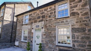 Un bâtiment en pierre avec une porte blanche, deux plantes en pot et un banc à l’extérieur.