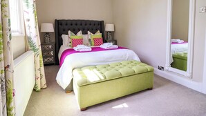 Un dormitorio con cama, lámparas de noche, un reposapiés verde, un radiador y una ventana con cortinas florales.