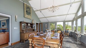 Uma sala de jantar com uma grande mesa de madeira posta para seis pessoas, cercada por cadeiras. Há um lustre sobre a mesa e vista para o exterior através de grandes janelas.