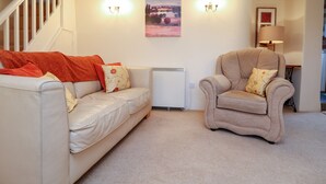 Una sala de estar con un sofá y un sillón color beige, y un radiador. Se ve una escalera a la izquierda y una lámpara a la derecha.
