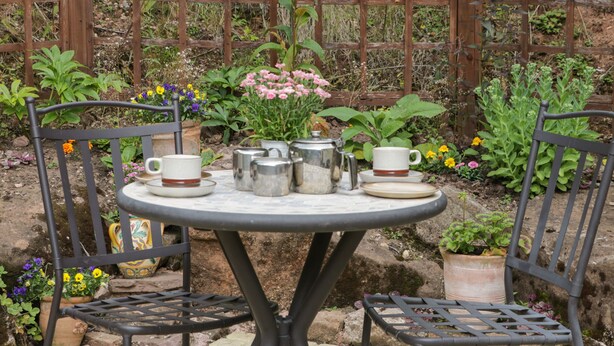 A round table with a metal frame, surrounded by two metal chairs. The table is set with a teapot, cups, and saucers. - Woods Edge (Mitcheldean)