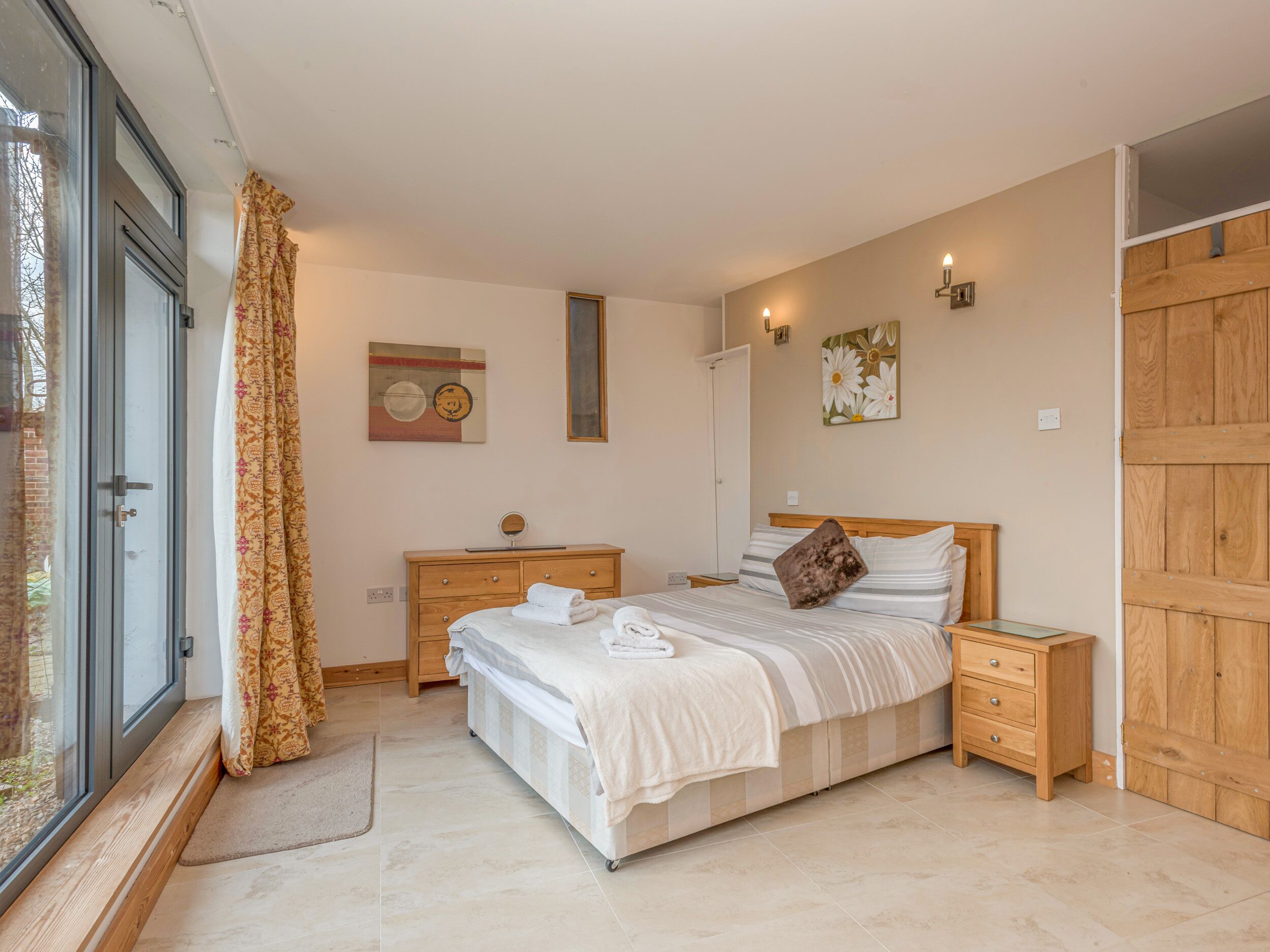 Een slaapkamer met een bed, houten nachtkastjes, een dressoir, een raam met gordijnen en een deur.