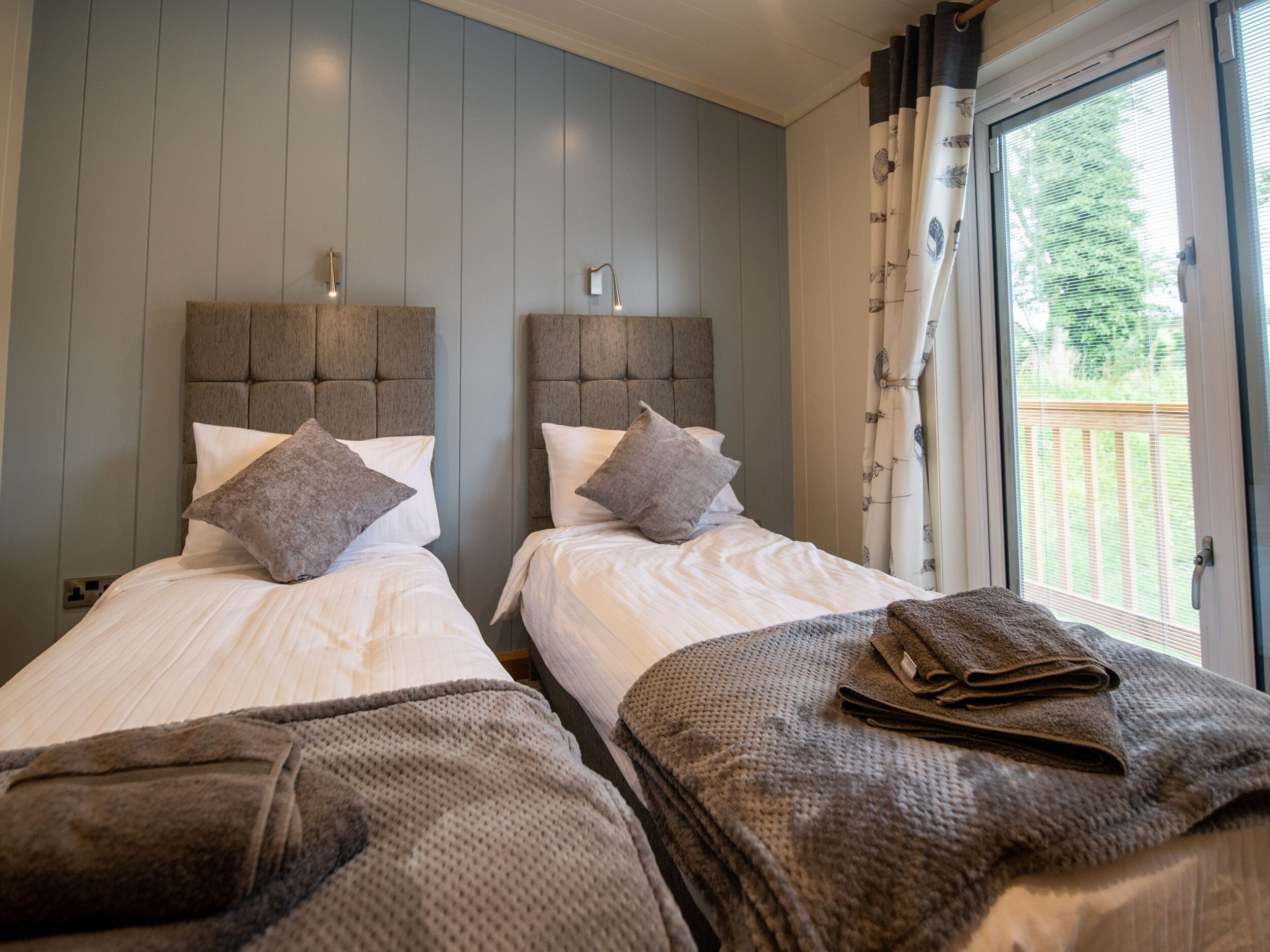 A hotel room with two beds, grey headboards, white bedding, and a view of the outdoors through a window with curtains.