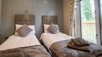 Ein Hotelzimmer mit zwei Betten, grauen Kopfteilen, weißer Bettwäsche und Blick ins Freie durch ein Fenster mit Vorhängen.