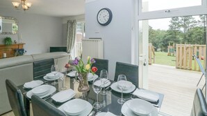 Un coin repas avec une table dressée pour quatre personnes, une horloge au mur et une vue sur un jardin par une porte ouverte.