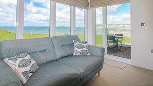 A room with a grey sofa, patterned cushions, a large window with a view of the sea, and a clock on the wall. - 7 Ocean's Edge (Kingsbridge)