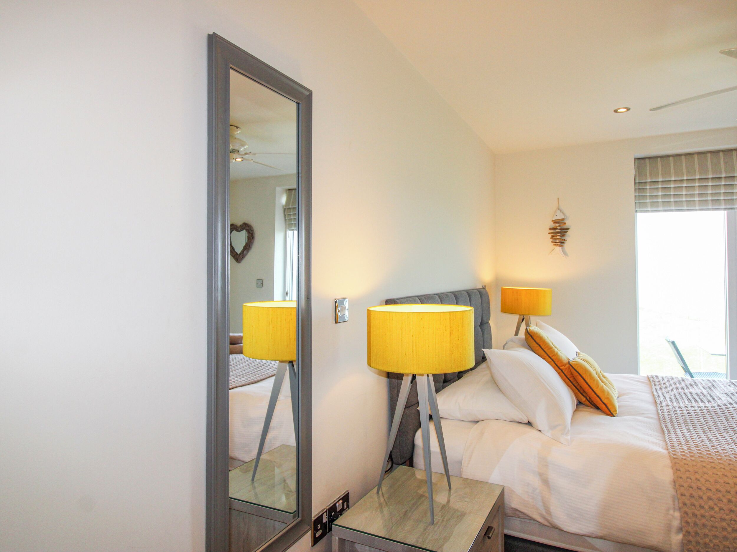 A hotel room with a bed, two bedside lamps, a mirror, a window with blinds, and a wall-mounted light fixture.