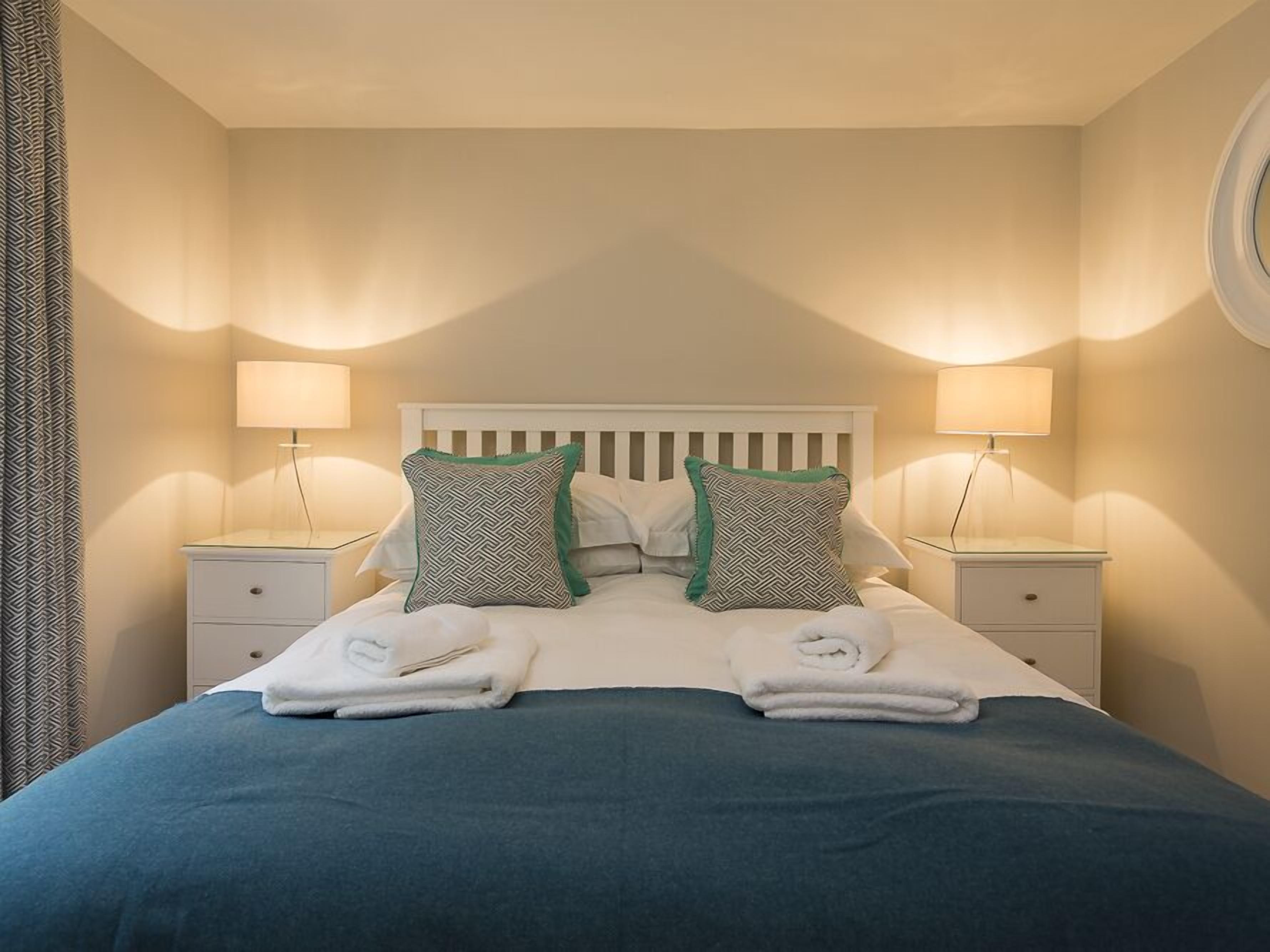 Cama bem arrumada com duas almofadas, duas mesas de cabeceira com luminárias e uma janela com cortinas.