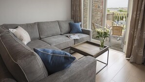 A modern living room with a grey sectional sofa, a coffee table, and a view of a stone building through a glass door. - 4 Keeper's Cottage, Hillfield Village (Dartmouth)