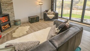 A modern living room with a stone wall, a fireplace, a leather sofa, and a large window with a view of the countryside. - Briar Barn (Bishop Auckland)
