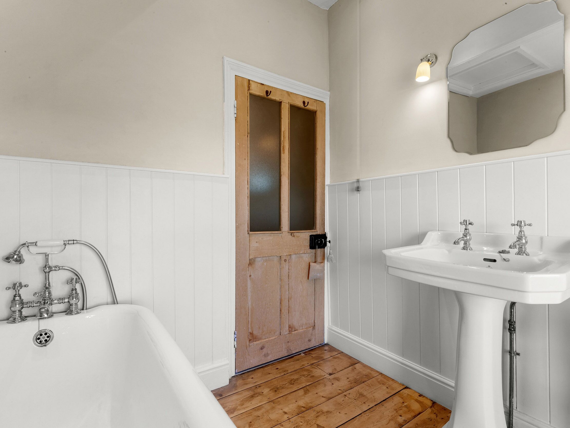 Une salle de bain avec une porte en bois, des lambris blancs, une baignoire blanche et un lavabo blanc doté de deux robinets.