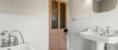 A bathroom with a wooden door, white wainscoting, a white bathtub, and a white sink with two faucets.