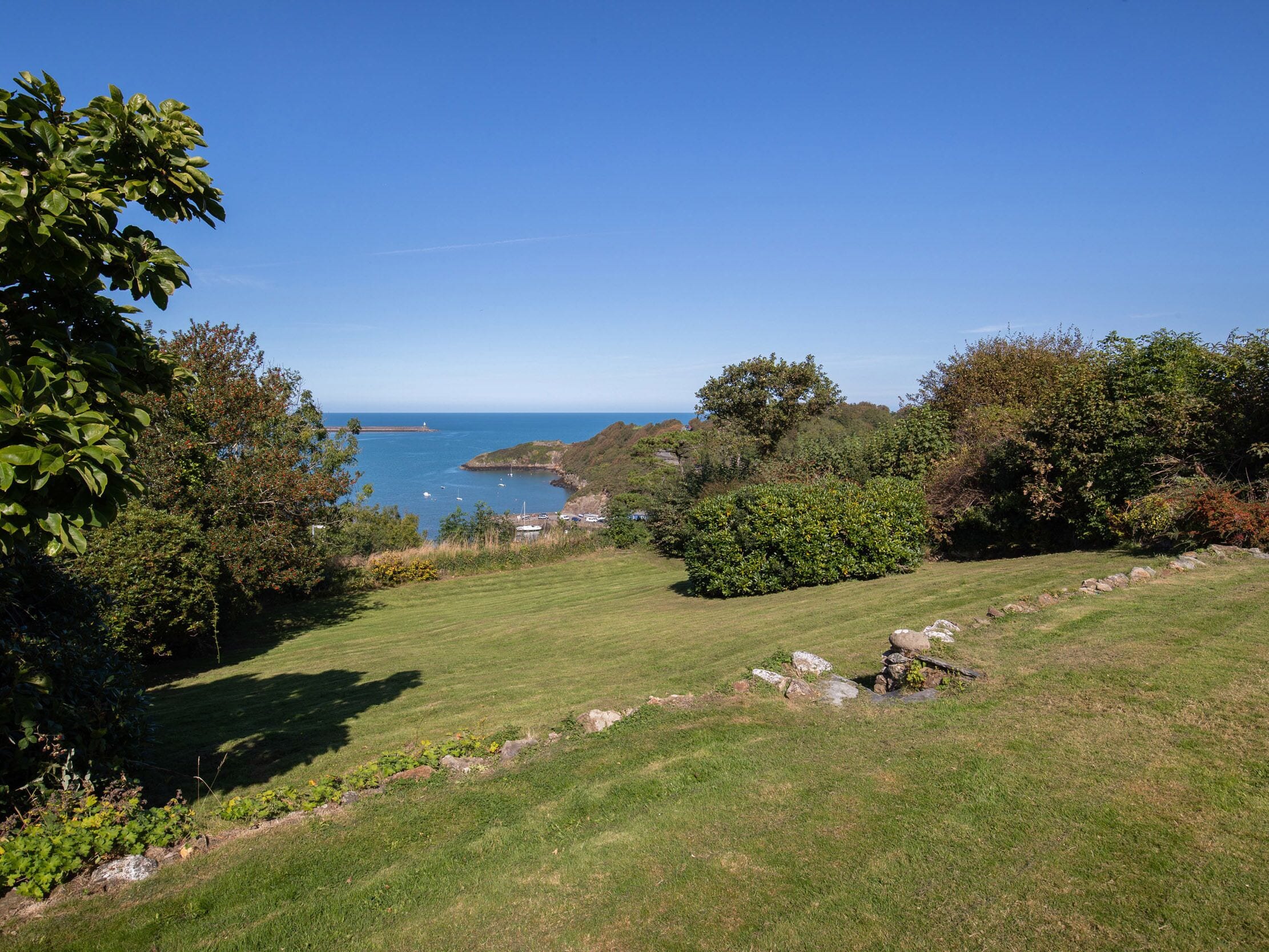 Eine Küstenlandschaft mit einer grasbewachsenen Fläche, Bäumen und Blick auf das Meer mit Booten.