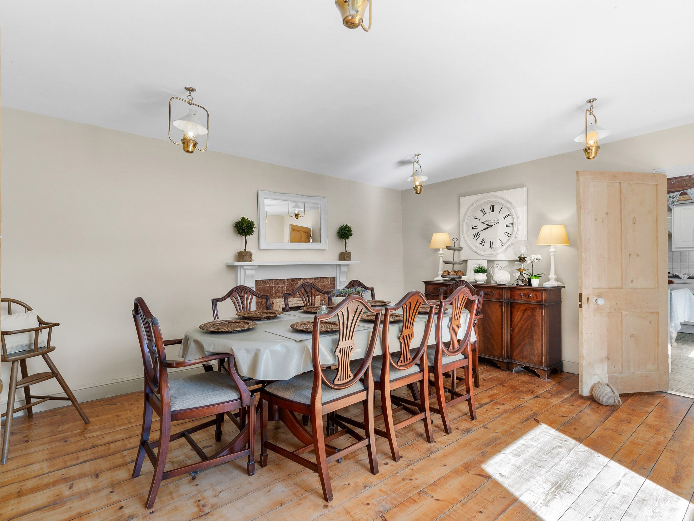 Une salle à manger avec une table en bois, six chaises, une cheminée, une grande horloge murale et deux lampes fixées au mur.