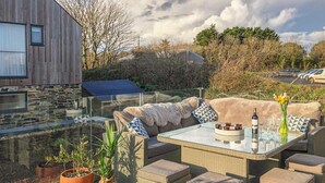 An outdoor patio with wicker furniture, a glass table, and a view of a stone building. - Idlewild, 2 Wheal Turnavore (St. Agnes)