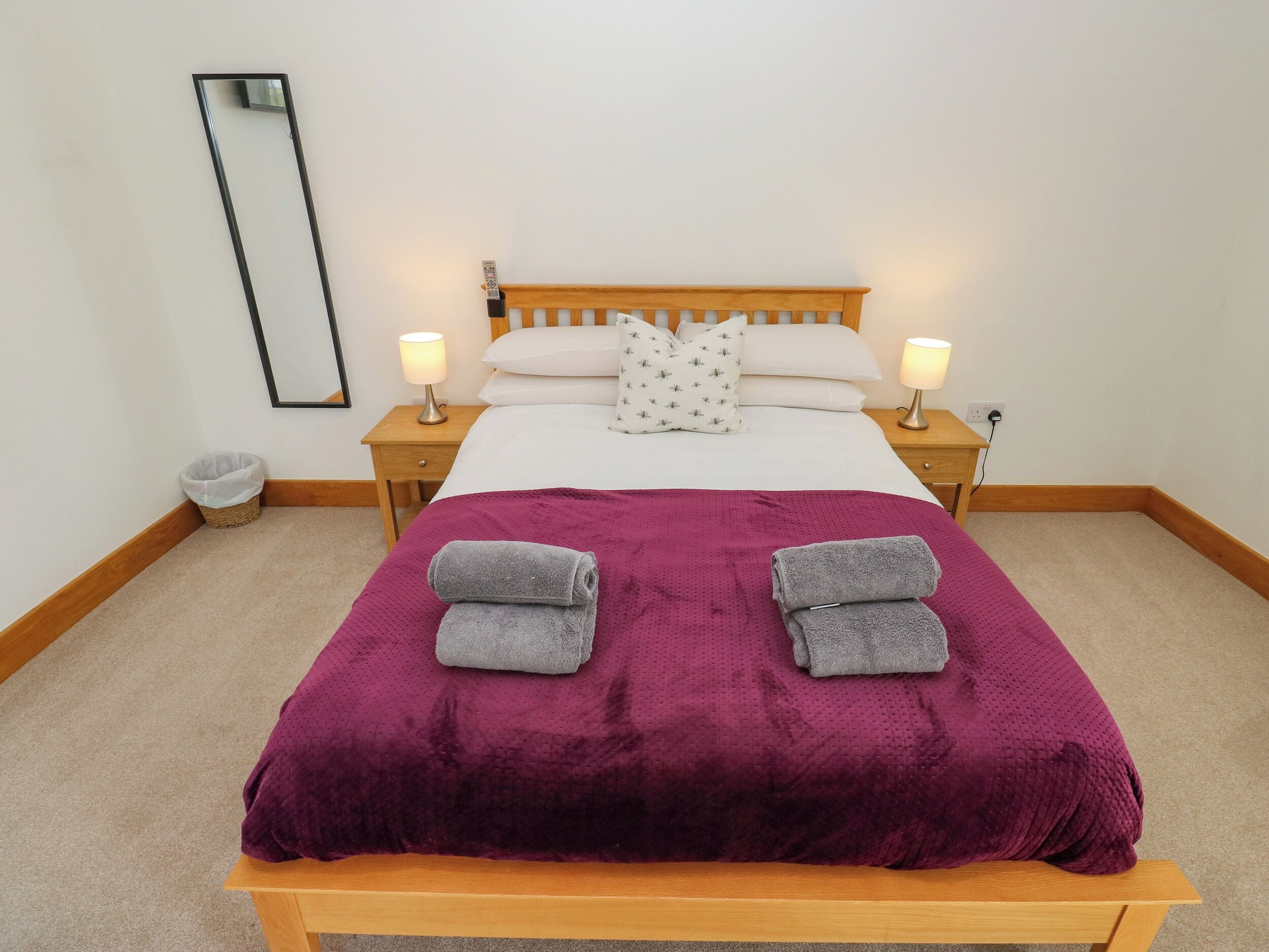 Una cama bien hecha con un edredón morado, dos almohadas y dos toallas enrolladas. Hay dos mesitas de noche con lámparas, un espejo en la pared y un cesto de basura pequeño.
