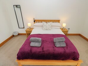 Una cama bien hecha con un edredón morado, dos almohadas y dos toallas enrolladas. Hay dos mesitas de noche con lámparas, un espejo en la pared y un cesto de basura pequeño.