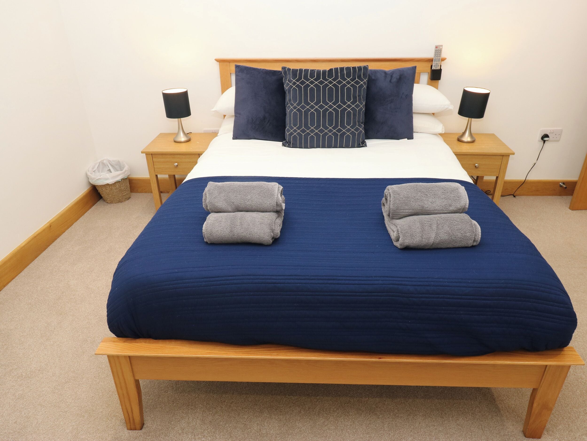 A neatly made bed with a blue bedspread, two pillows, and two folded towels. There are two bedside tables with lamps and a small trash bin.