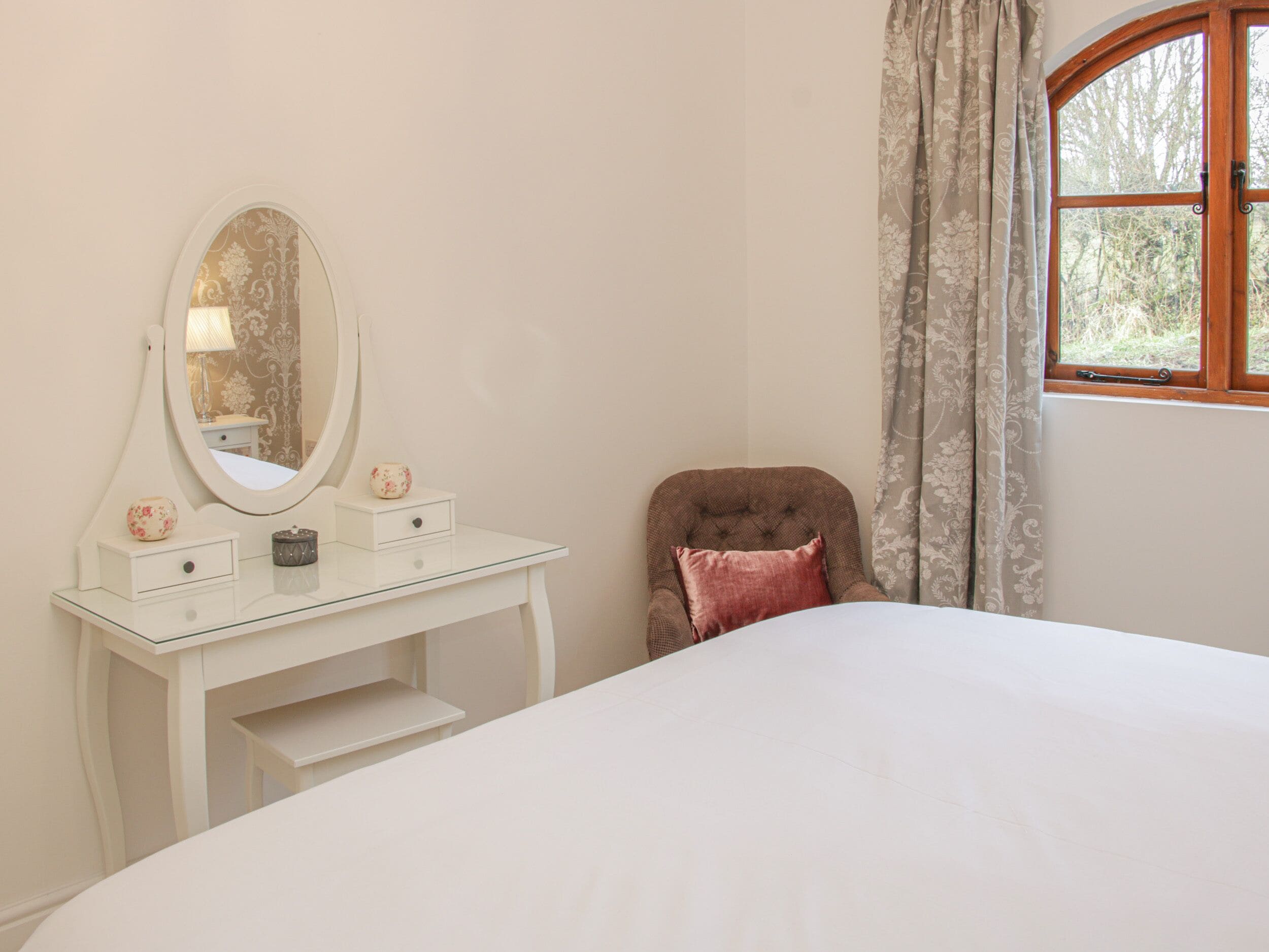Une chambre à coucher avec un lit blanc, une coiffeuse avec un miroir, une chaise et une fenêtre avec des rideaux.