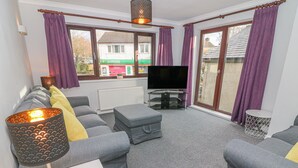 A living room with grey sofas, a TV on a stand, a lamp, and a view of a building with a sign reading "LONDIS". - Rose Cottage (Grange-over-Sands)