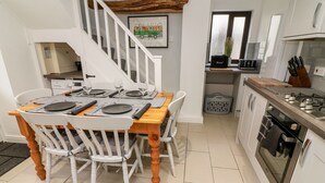 A dining area with a wooden table set for four, white chairs, and a staircase leading to another room. - Rose Cottage (Grange-over-Sands)