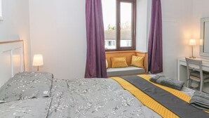 A bedroom with a bed, a window with curtains, a desk with a lamp, and a view of the outside. - Rose Cottage (Grange-over-Sands)