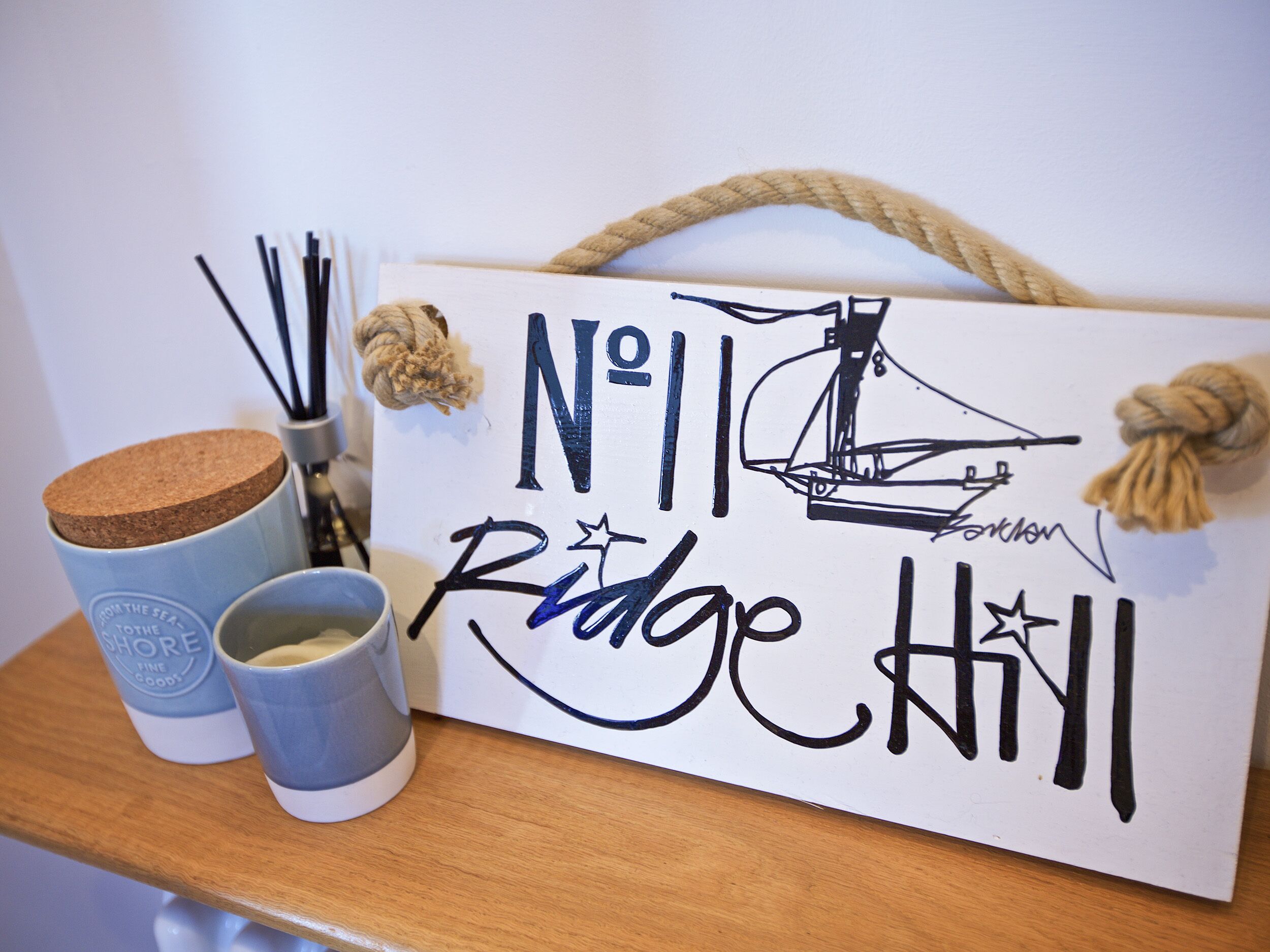 Un cartel con la inscripción 'No 11 Ridge Hill', una ilustración de un barco, un recipiente de corcho y dos tazas sobre una superficie de madera.