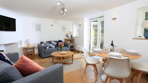 Uma sala de estar moderna com um sofá cinzento, uma mesa de centro e um espaço de jantar com cadeiras brancas e uma mesa de madeira.