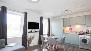 A modern apartment with a living room, dining area, and kitchen. It includes a flat-screen TV, a dining table with chairs, a sofa, a glass coffee table, a washing machine, and a radiator. - The Nook (Inverness)