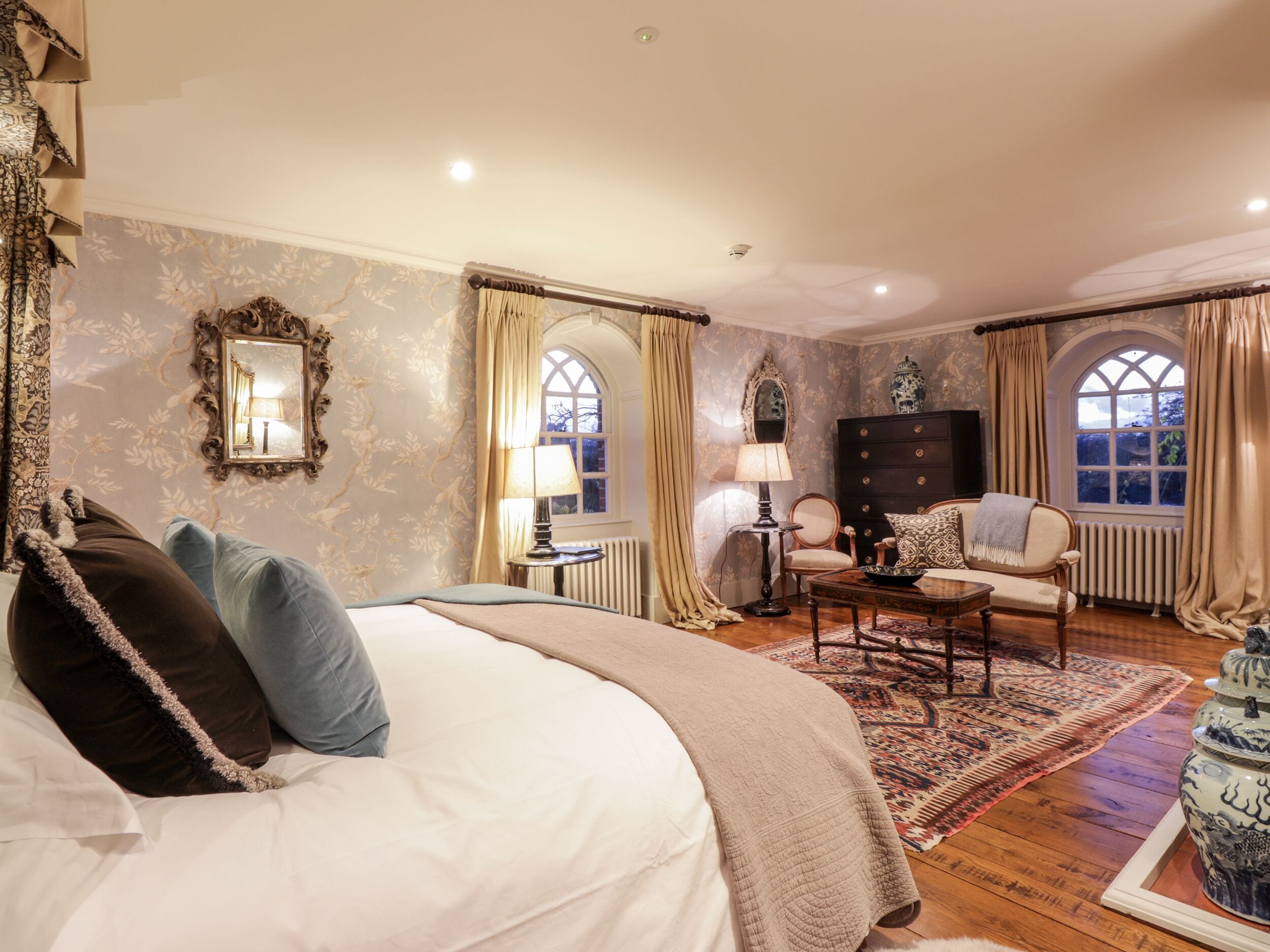 A bedroom with a large bed, a dresser, a chair, and a table. There are two windows with curtains, a mirror, and a radiator.