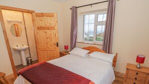 Un dormitorio con cama de madera, una mesita de noche con lámpara, una ventana con cortinas, una puerta de madera y un baño con lavamanos y espejo.
