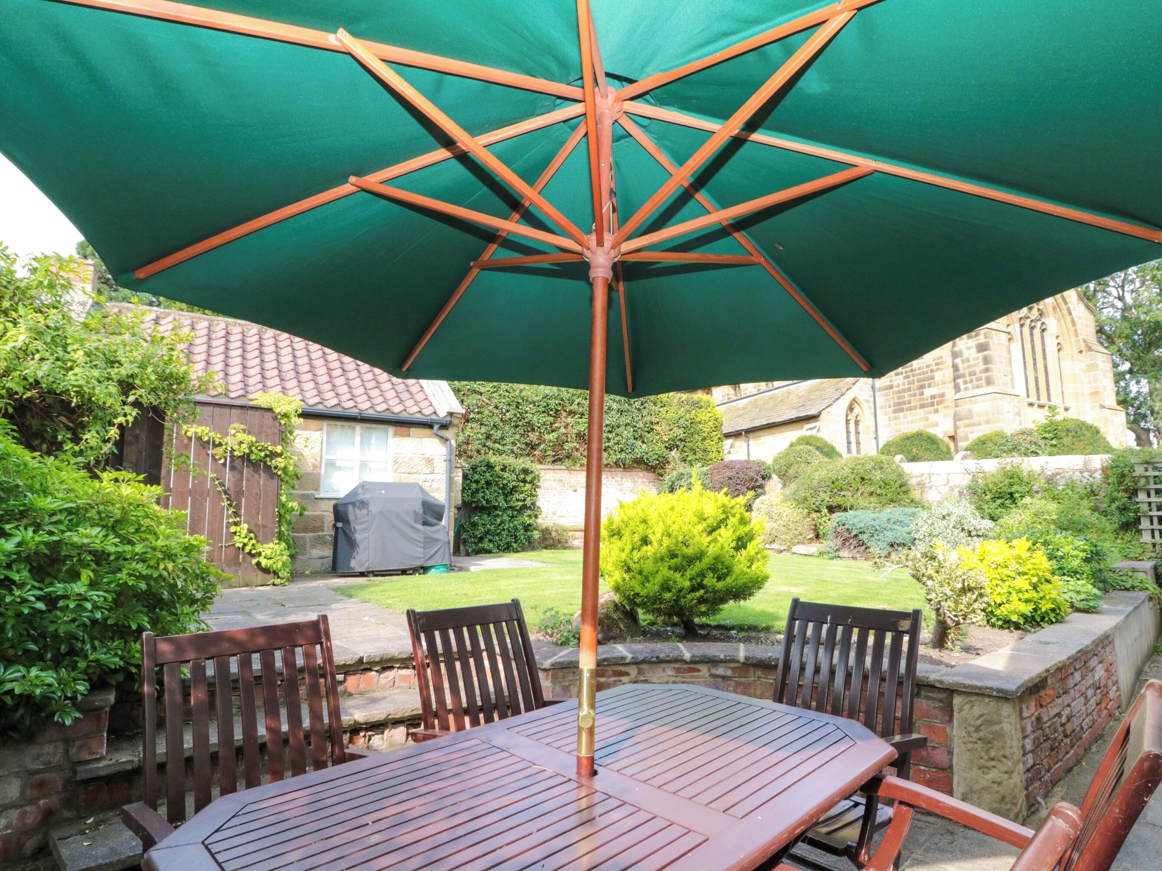 A patio with a wooden table and chairs, a green umbrella, and a stone wall.
