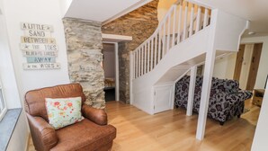 Un vaste espace de vie comprenant un fauteuil en cuir marron, un canapé à motifs et un escalier blanc. On peut également apercevoir un mur en pierre et un plafond en bois.