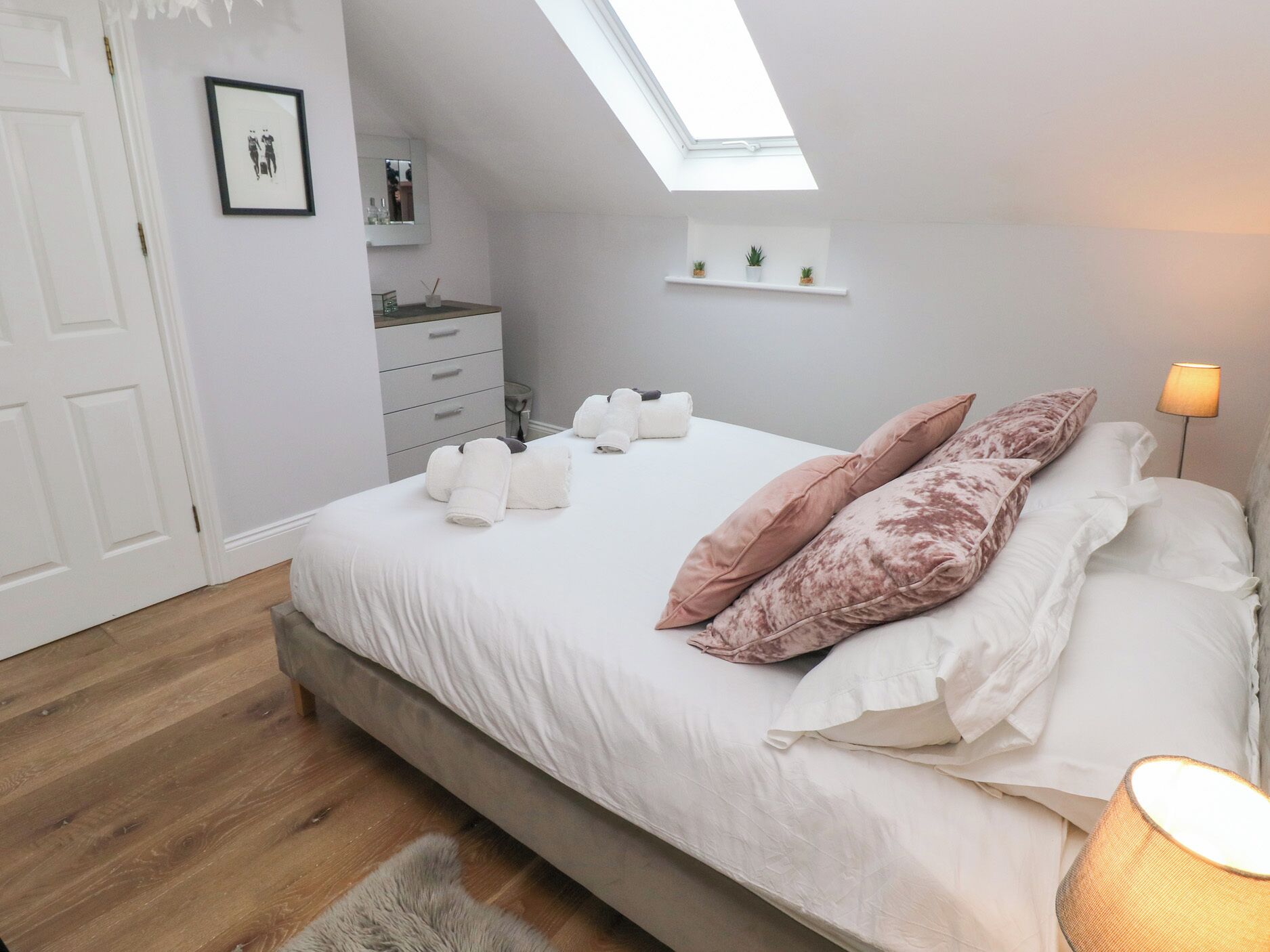 Une chambre avec une lucarne, un lit recouvert de draps blancs et agrémenté d’oreillers roses, une table de chevet avec des articles de toilette et une petite étagère avec des plantes.