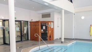 An indoor swimming pool with clear blue water, a wooden sauna, and a glass door entrance. - Cambrie (Tenby)