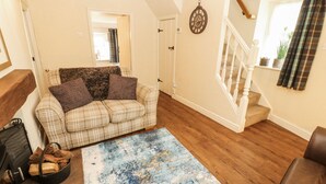 A cozy living room with a plaid sofa, a blue abstract rug, and a fireplace with logs. - Corner Cottage (Carnforth)
