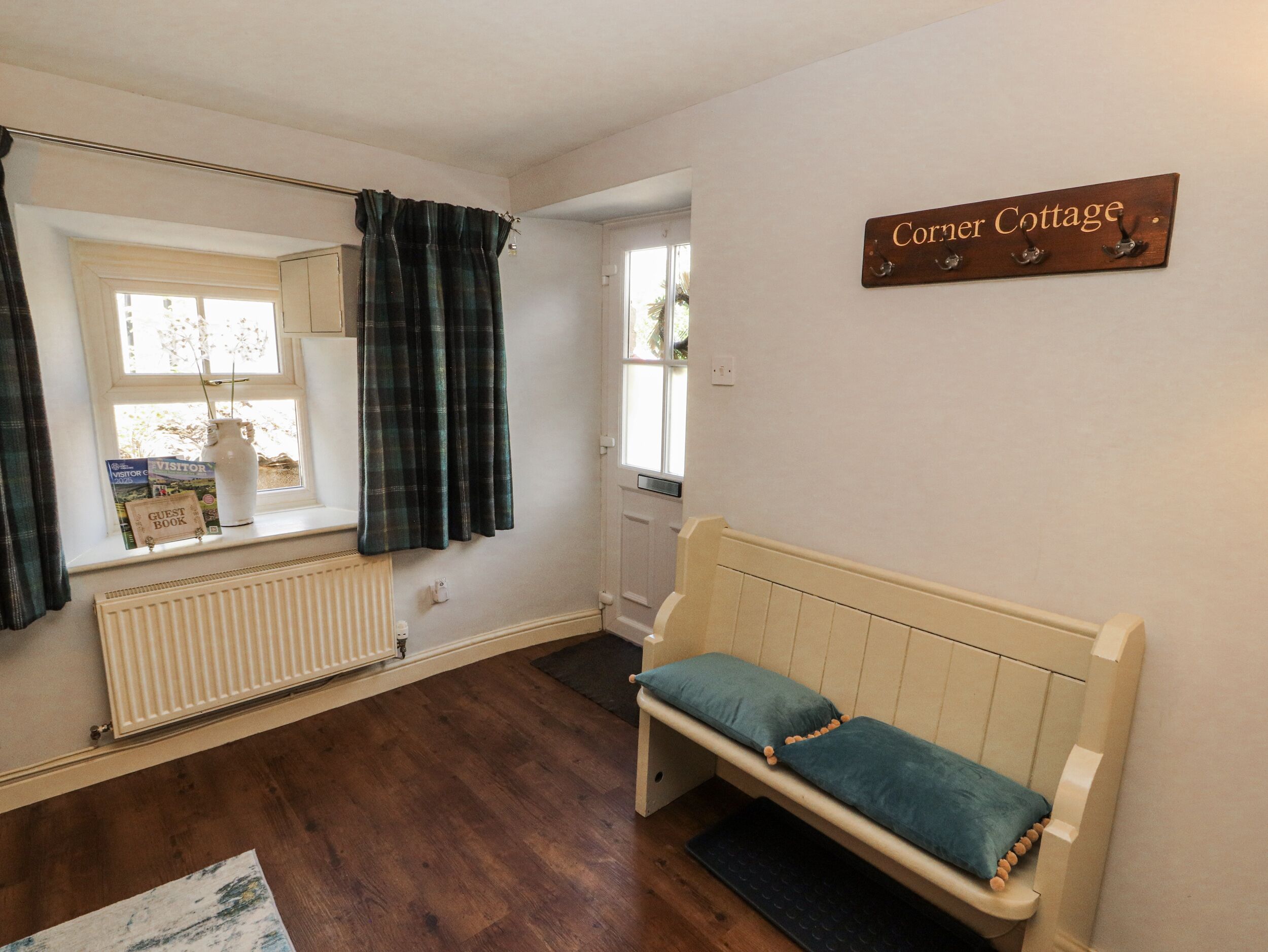 A cozy room with a wooden bench, a radiator, a window with curtains, and a sign reading "Corner Cottage".