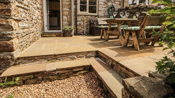 A stone building with a patio area featuring a wooden table and chairs, a small garden, and a stone pathway leading to the entrance. - Corner Cottage (Carnforth)