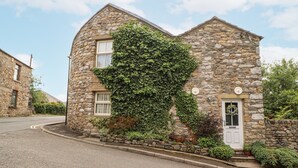 A stone house with ivy covering part of the facade, a white door, and a small garden area with potted plants. - Corner Cottage (Carnforth)