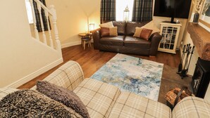 A cozy living room with a brown leather sofa, plaid cushions, a small side table, a bookshelf, and a fireplace. - Corner Cottage (Carnforth)