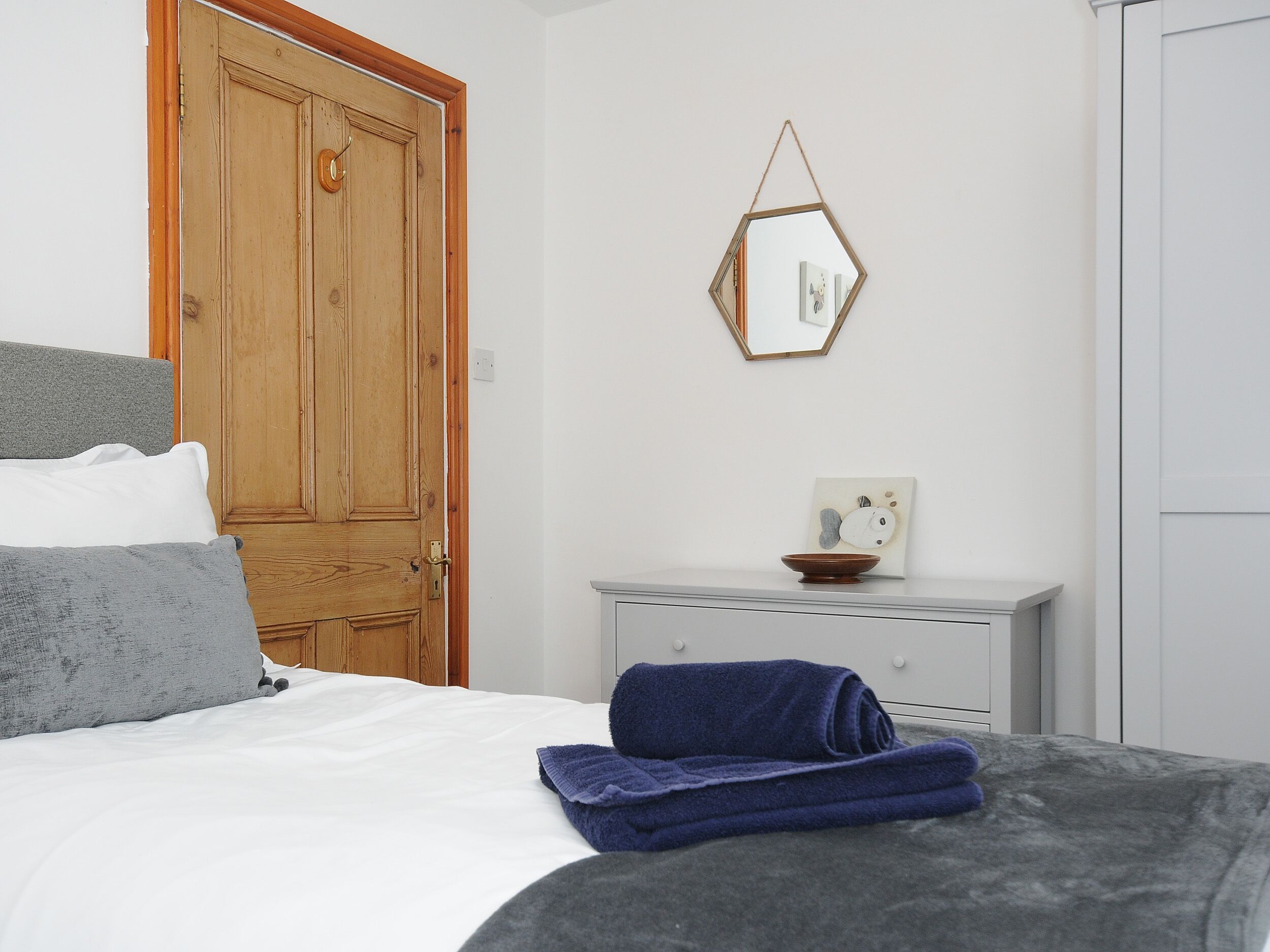 A bedroom with a wooden door, a bed with white and grey bedding, a blue towel, a small white dresser, and a hexagonal mirror on the wall.
