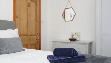 A bedroom with a wooden door, a bed with white and grey bedding, a blue towel, a small white dresser, and a hexagonal mirror on the wall.