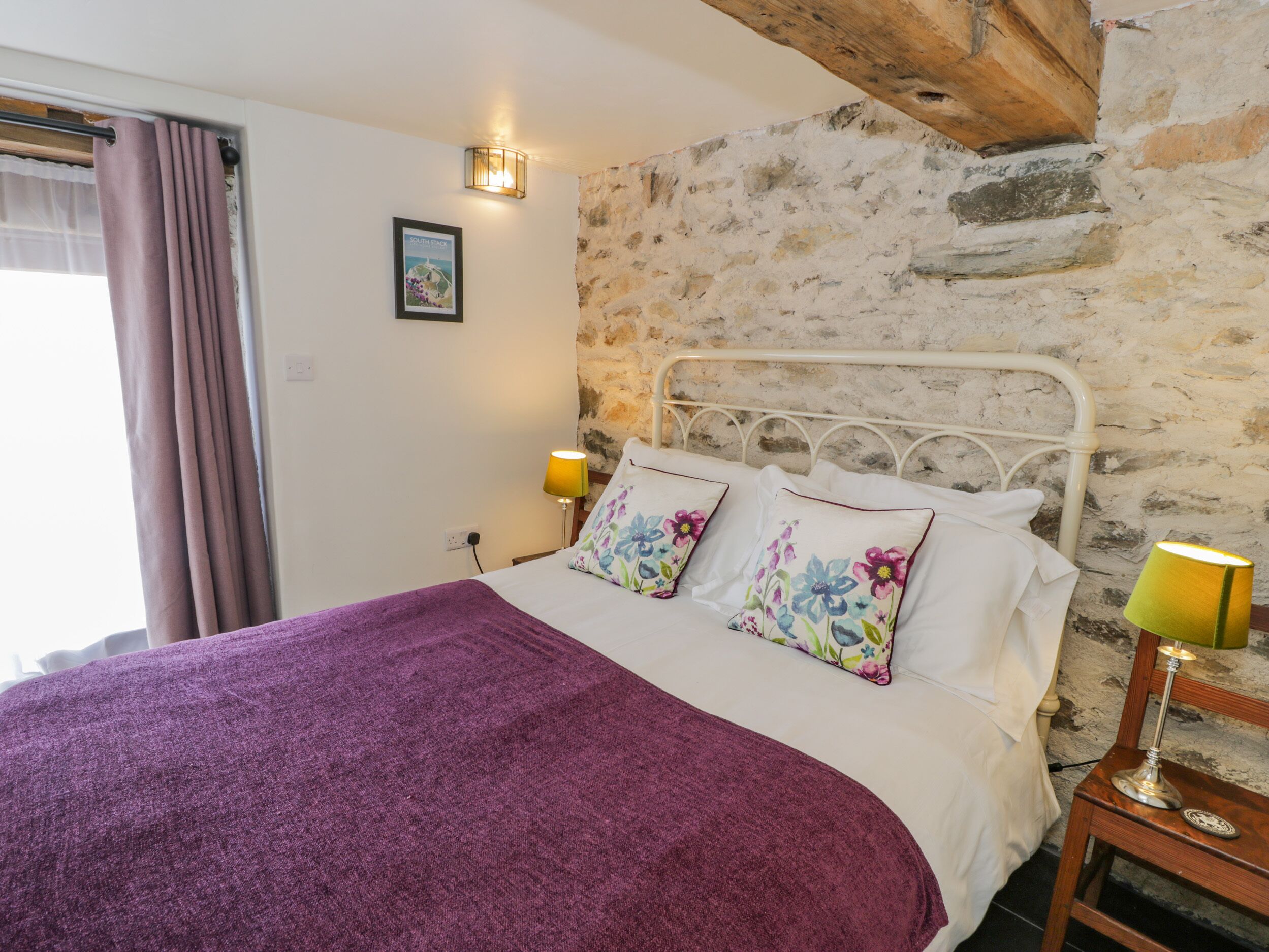 A bed with a purple blanket, floral pillows, and a white bedspread. There are two bedside lamps and a picture on the wall.
