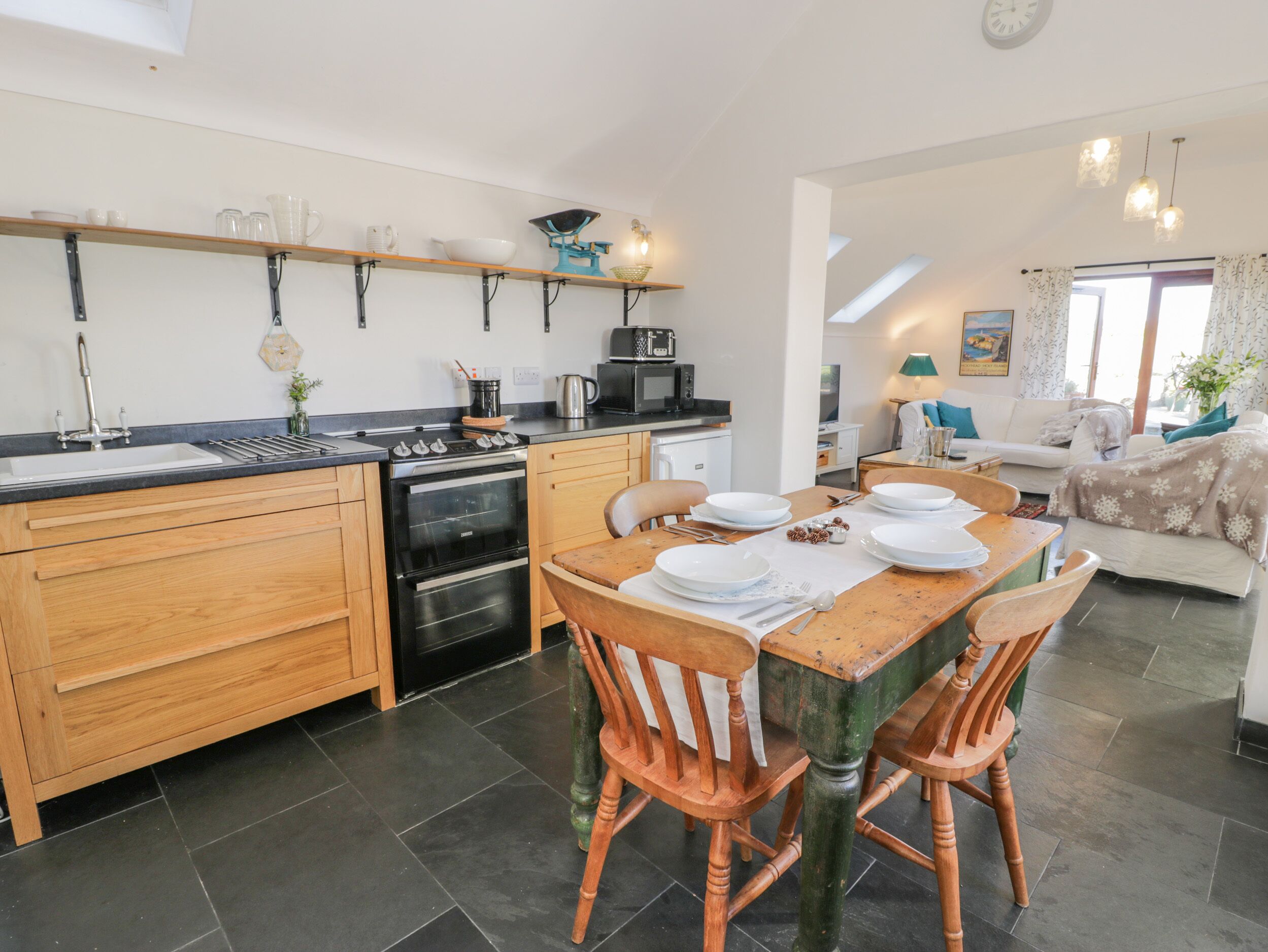 A modern kitchen with wooden cabinets, a dining table set for four, and a living area with a sofa and a TV.