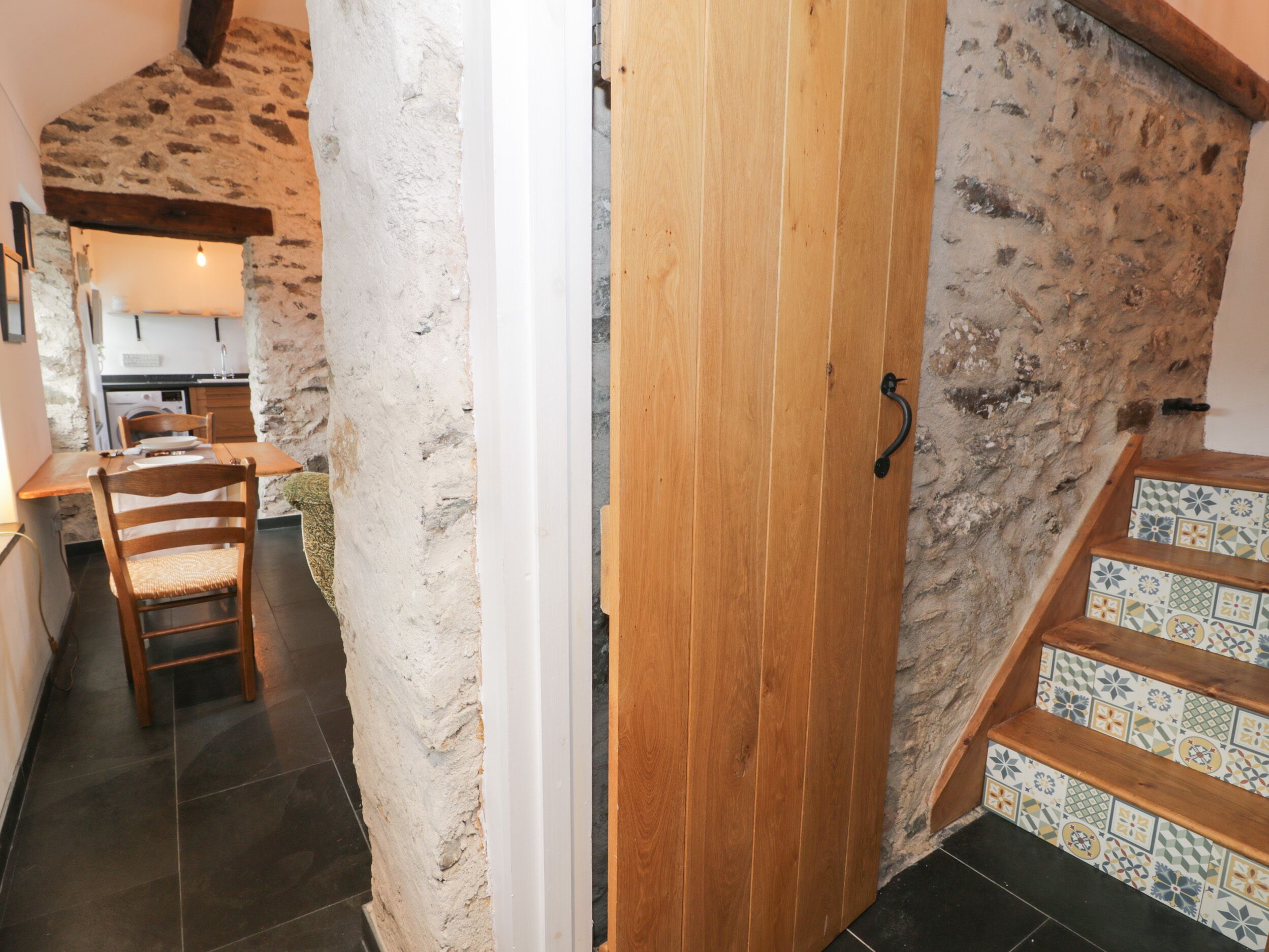 A narrow hallway with a wooden door, a dining area with a table and chairs, and a staircase with patterned tiles.