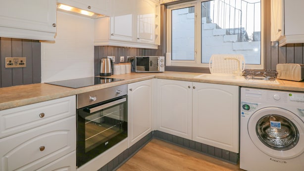 Una cocina compacta con armarios blancos, horno integrado, microondas, lavadora y ventana con vistas a una valla blanca.