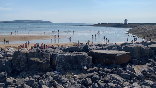 A rocky beach with people enjoying the water and the view of a lighthouse in the distance.